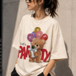 Woman wearing Luri Let's Party graphic oversized t-shirt in cream — front print tee with fluffy teddy bear holding colourful balloons and champagne bucket, bold red "Let's PARTY" text, unisex oversized fit