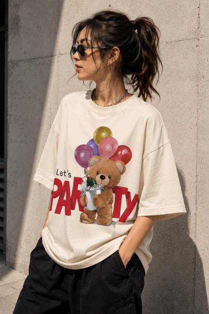 Woman wearing Luri Let's Party graphic oversized t-shirt in cream — front print tee with fluffy teddy bear holding colourful balloons and champagne bucket, bold red "Let's PARTY" text, unisex oversized fit